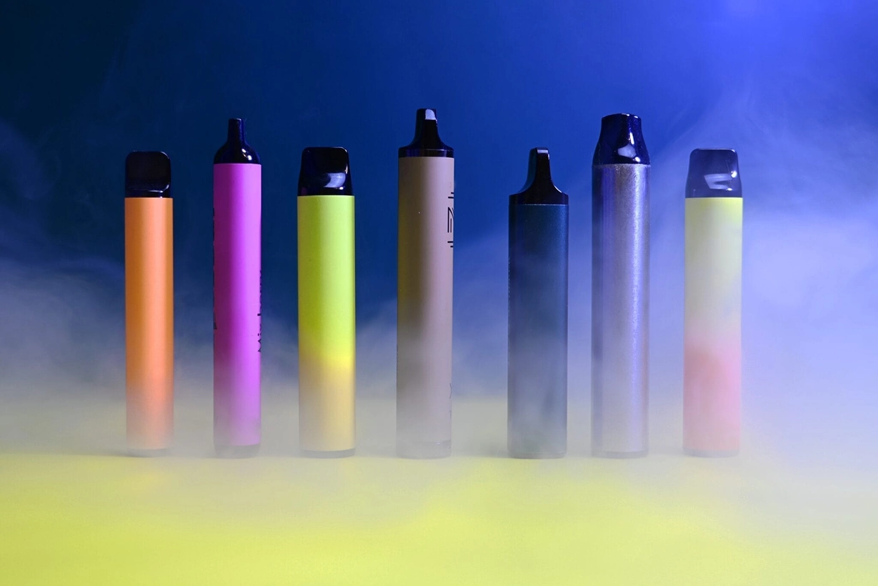 Une rangée de sept cigarettes électroniques jetables (puffs) colorées, alignées verticalement au milieu d'un nuage de fumée ou de vapeur dense