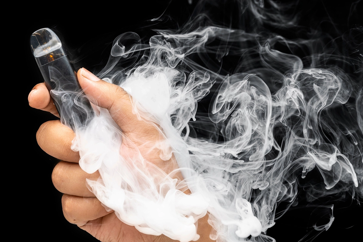 Une main tenant une cigarette électronique noire et transparente, entourée de volutes épaisses de vapeur blanche sur un fond entièrement noir.